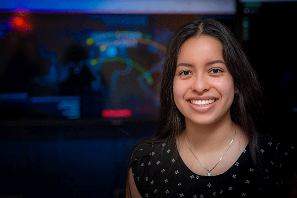 A young Latina woman with long dark hair smiles at the camera in a computer lab. The woman is wearing a black shirt with a white pattern and a silver necklace. A video monitor showing a colorful map of simulated cyberattacks is blurred but visible behind her.