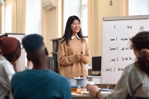 A smiling Asian woman with shoulder-length black hair stands in front of several peers in a classroom with a black marker in her hand. She wears a white collared shirt underneath a tan sweater with blue jeans. To her right is a whiteboard with what look like syllables of another language written out and English words – “I,” “I want,” “I need,” “I can,” “I think.” – written in black marker next to them. Her peers, blurred but visible in the foreground, include a Black man with short hair.